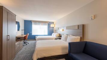 Quarto de hotel com duas camas, uma escrivaninha, uma cadeira, um sofá e uma janela grande.