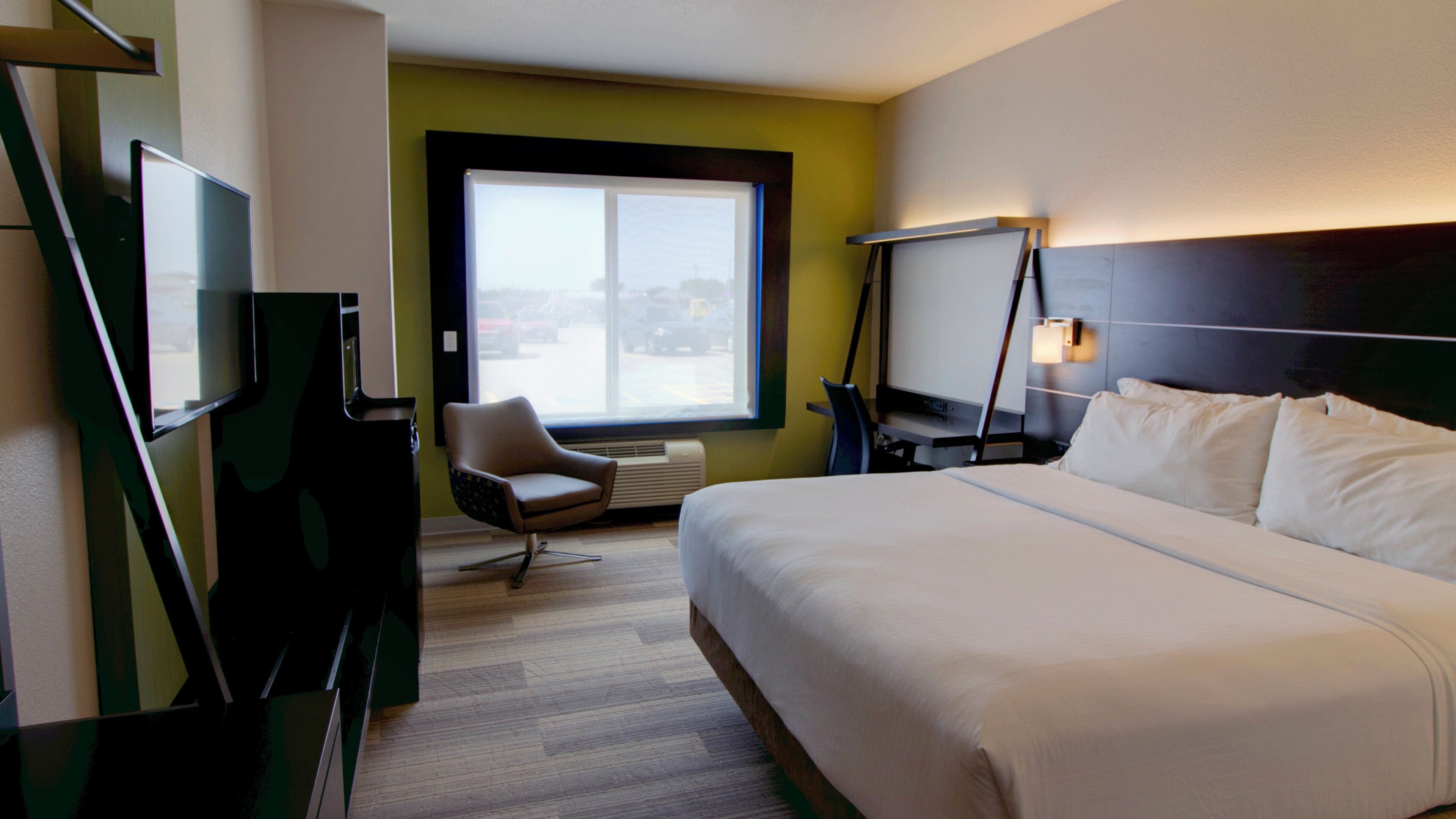 A hotel room with a large bed, a desk with a computer, a chair, a window with a view, and a flat-screen TV.