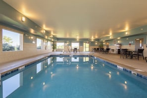 A swimming pool with clear water, surrounded by a lounge area with chairs and tables, and large windows providing natural light.