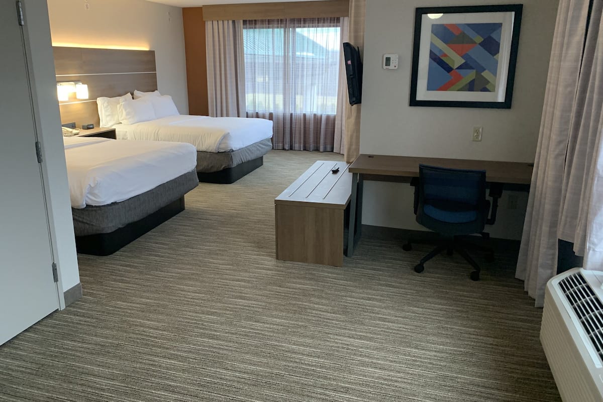 Une chambre d’hôtel avec deux lits, un bureau, une chaise, une petite table, un radiateur et une fenêtre avec des rideaux.