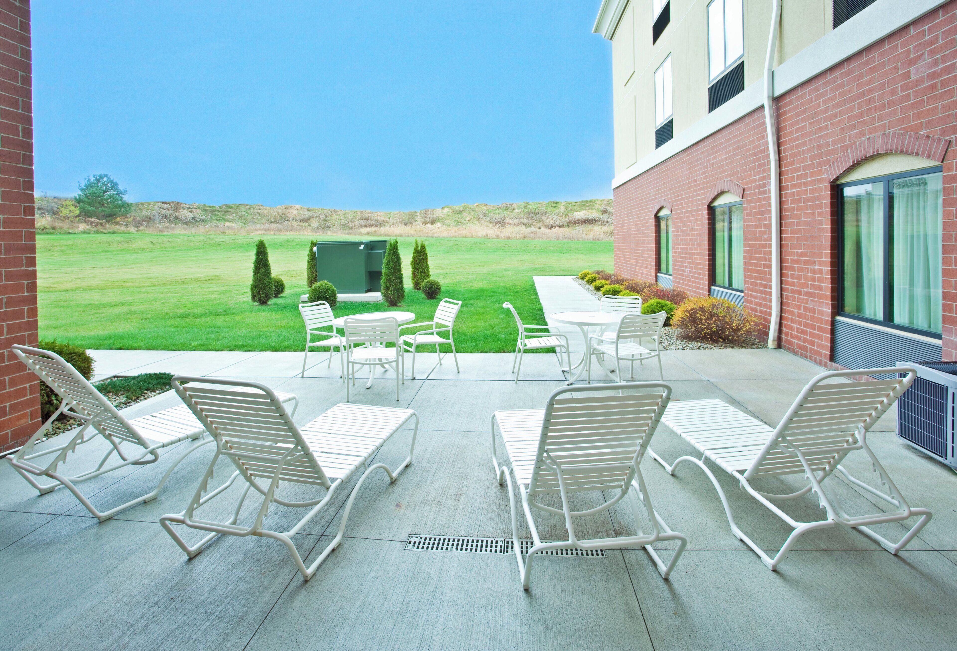 An outdoor patio with white metal chairs and tables, a grassy lawn, and a brick building in the background.