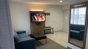 Een moderne hotelkamer met een flatscreen televisie aan de muur, een blauwe bank, een klein houten tafeltje en een spiegel die de kamer weerspiegelt.