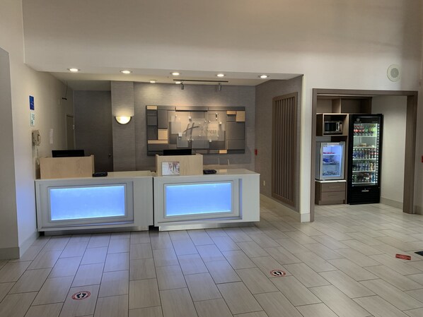Un espace d’accueil d’hôtel au design moderne, comprenant un comptoir d’accueil, un écran fixé au mur et un réfrigérateur.