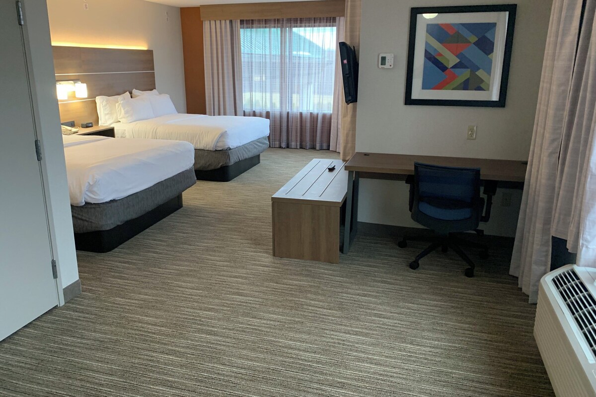 Ein Hotelzimmer mit zwei Betten, einem Schreibtisch, einem Stuhl, einem kleinen Tisch, einem Heizgerät und einem Fenster mit Vorhängen.