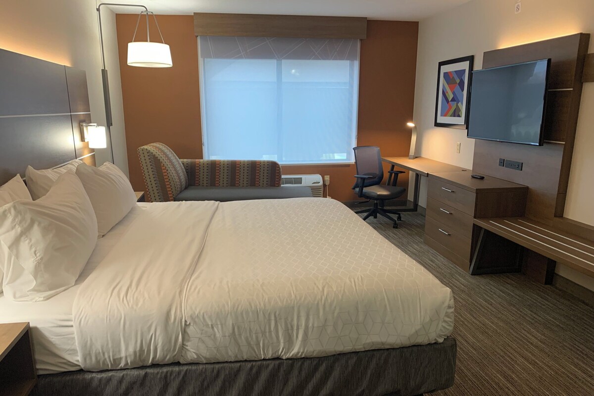 Ein Hotelzimmer mit einem großen Bett, einem Schreibtisch, einem Stuhl, einem Fernseher und einem Fenster mit Jalousien.