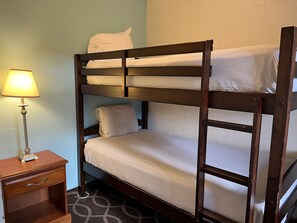 A bunk bed with white bedding, a bedside lamp, and a wooden nightstand with a drawer.