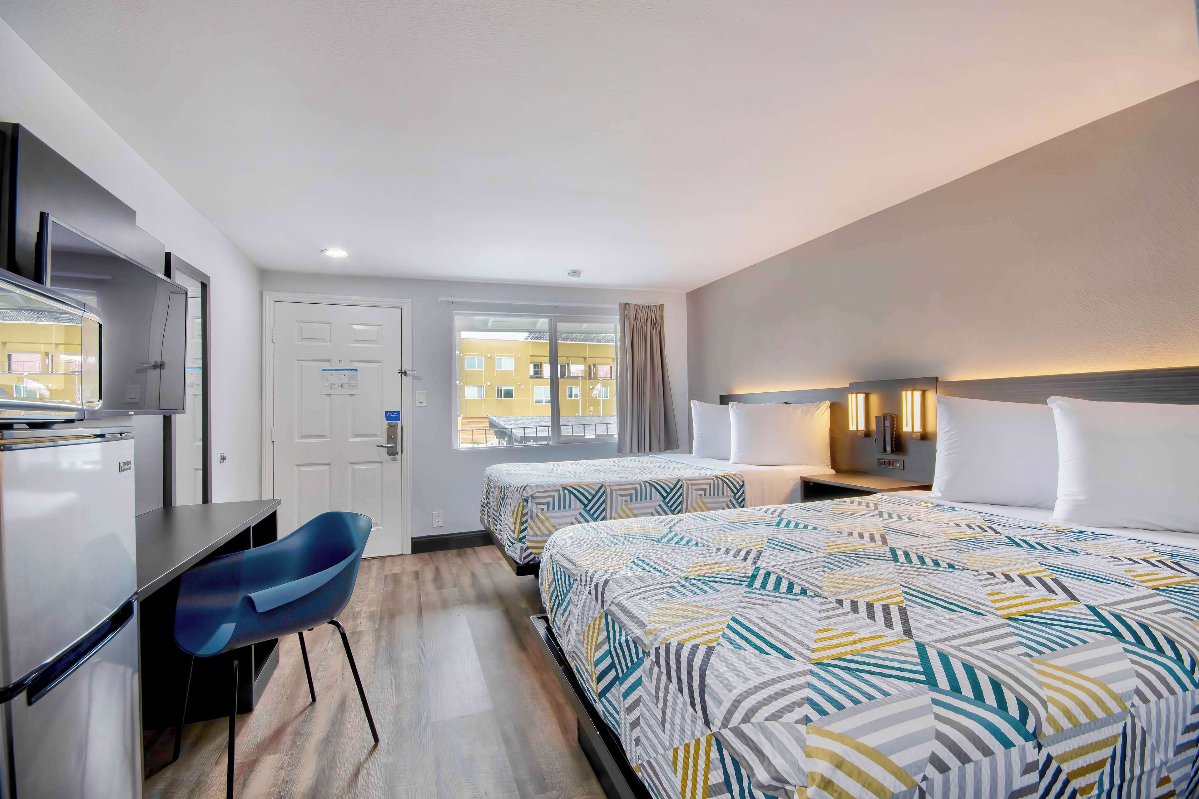 Quarto de hotel com duas camas, uma mesa, uma cadeira, uma televisão e uma janela com vista para edifícios.