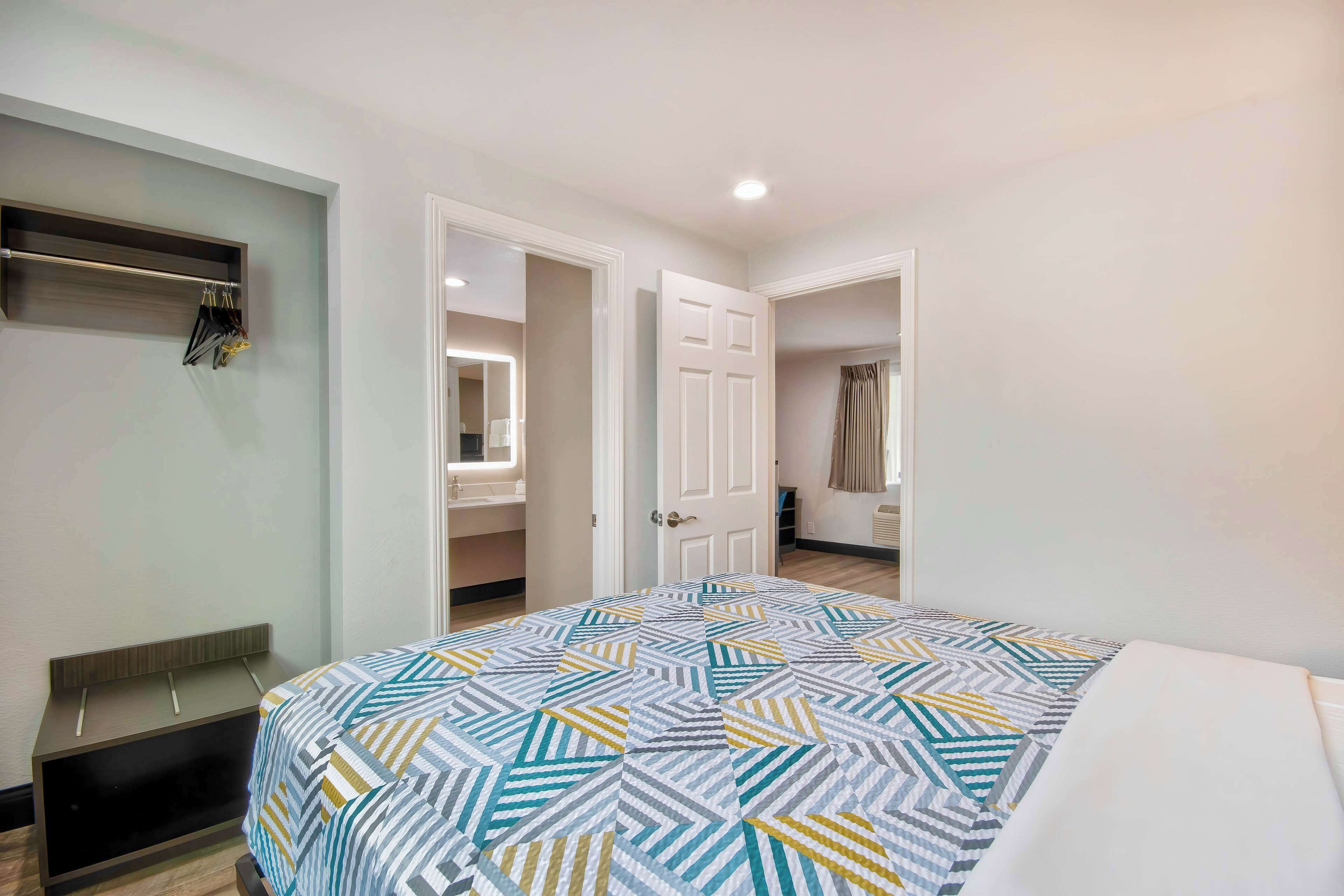 Un dormitorio con cama, mesita de noche, espejo y puerta que da a otra habitación.