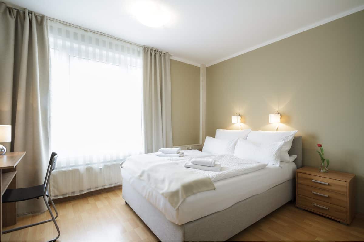 Ein ordentlich bezogenes Bett mit weißen Leinen, ein Nachttisch mit Lampe und ein Sessel.