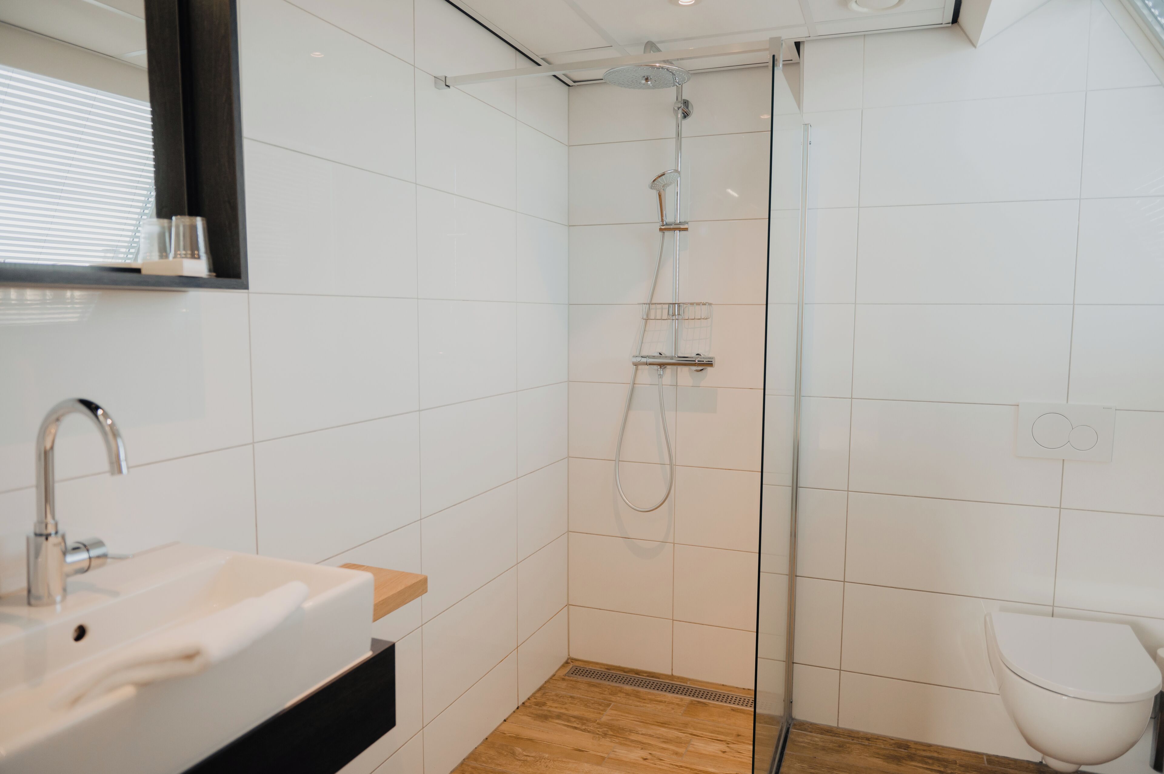Un baño moderno con ducha de vidrio, lavamanos blanco sobre encimera de madera y un inodoro blanco.
