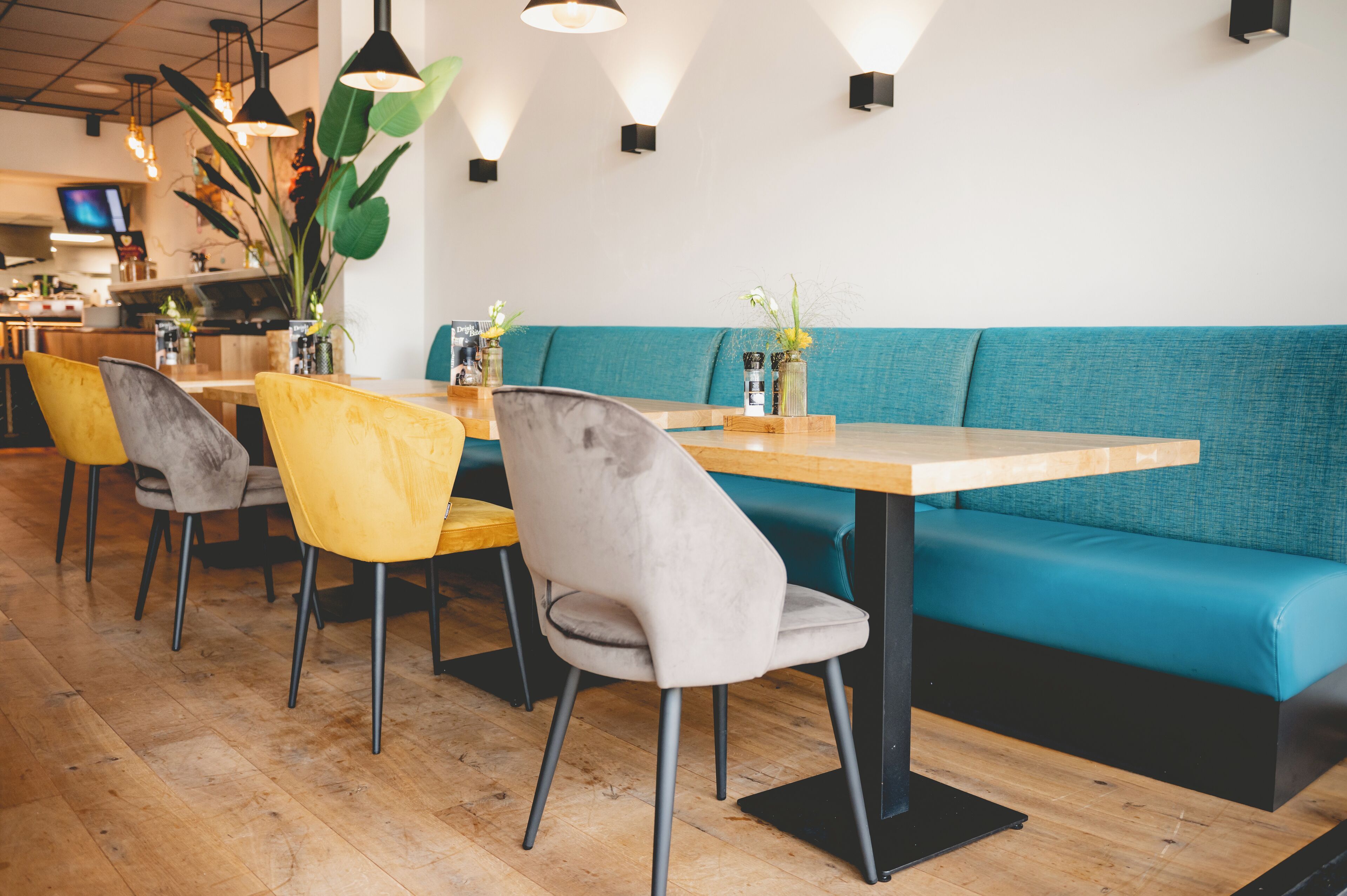 A restaurant interior with wooden tables, yellow and grey chairs, teal cushioned seating, and black pendant lighting.