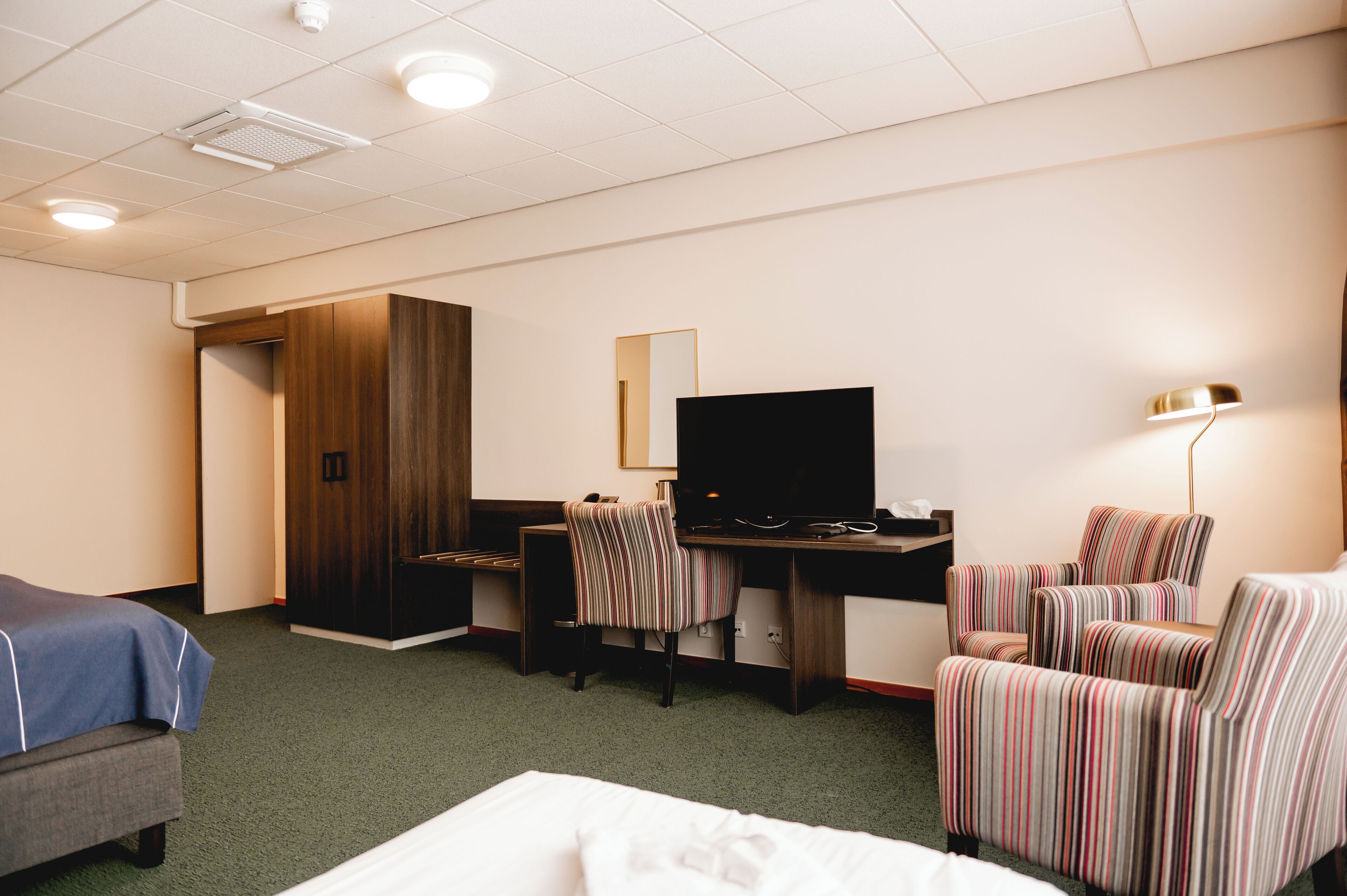 Habitación de hotel con una cama, un escritorio con computadora, un televisor, un sillón a rayas y una lámpara.