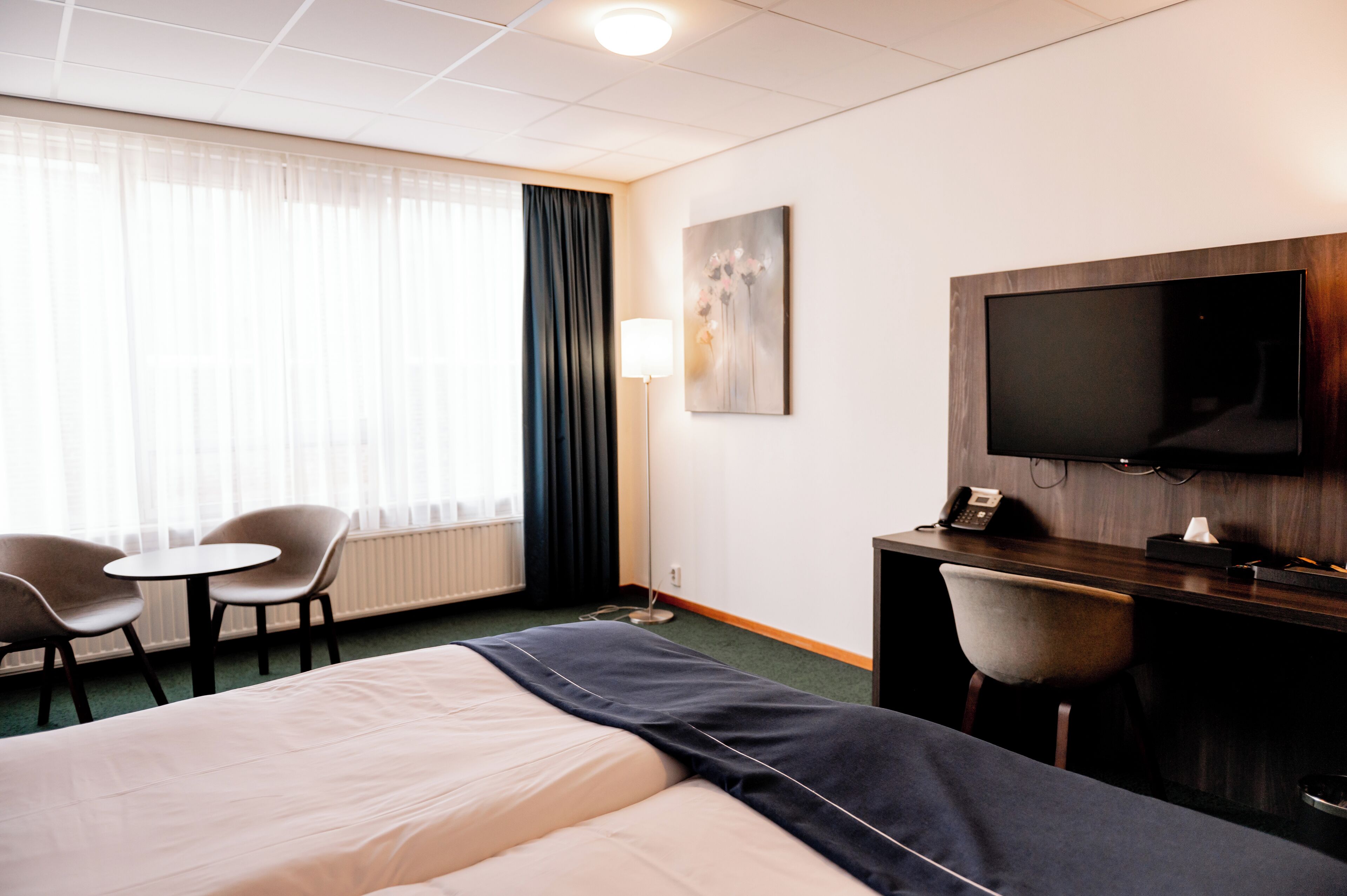 Habitación de hotel con cama, dos sillas, escritorio, televisor de pantalla plana, lámpara y ventana con cortinas.