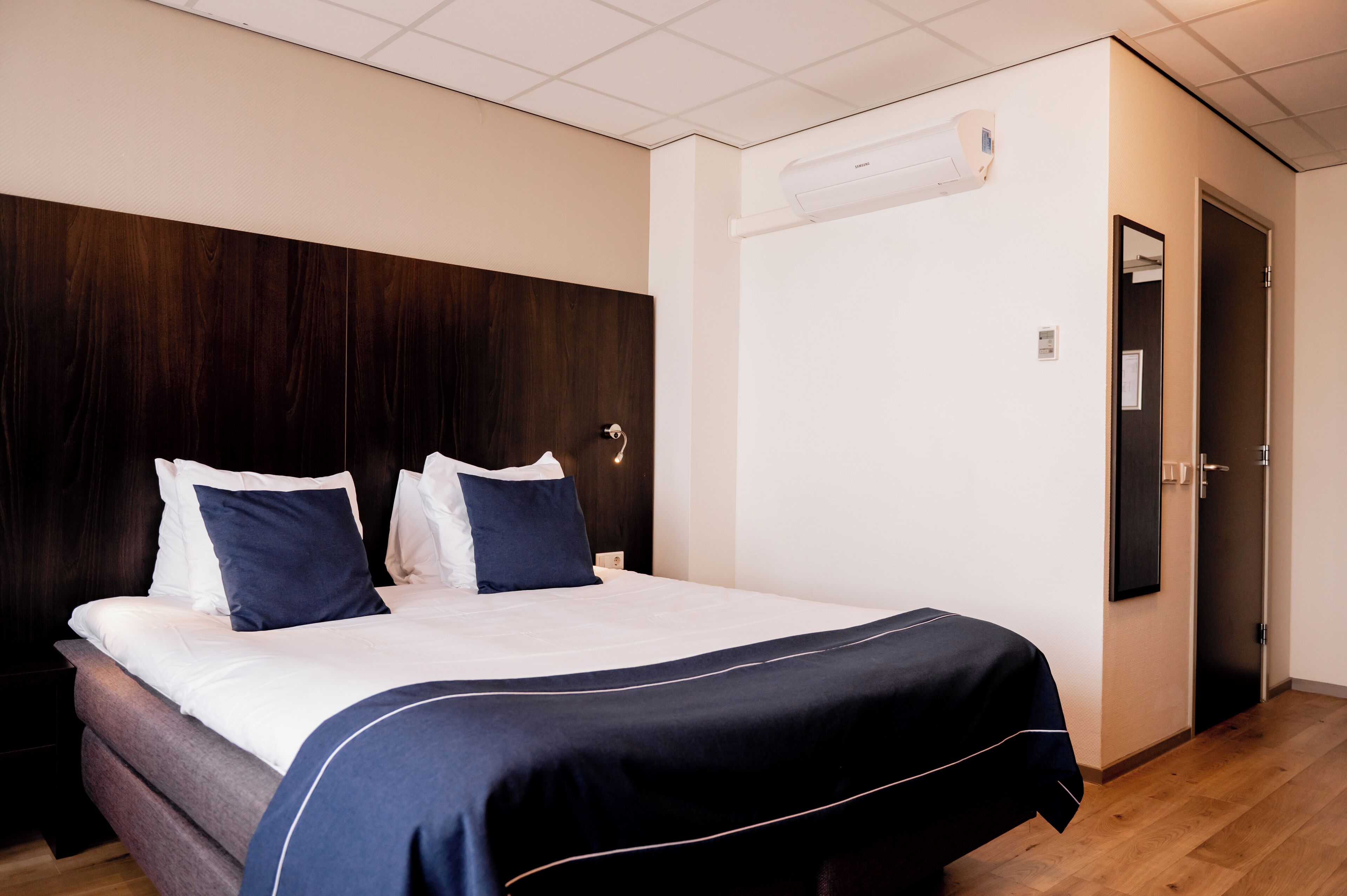 A hotel room with a bed, a dark wooden headboard, a white wall, and a door.