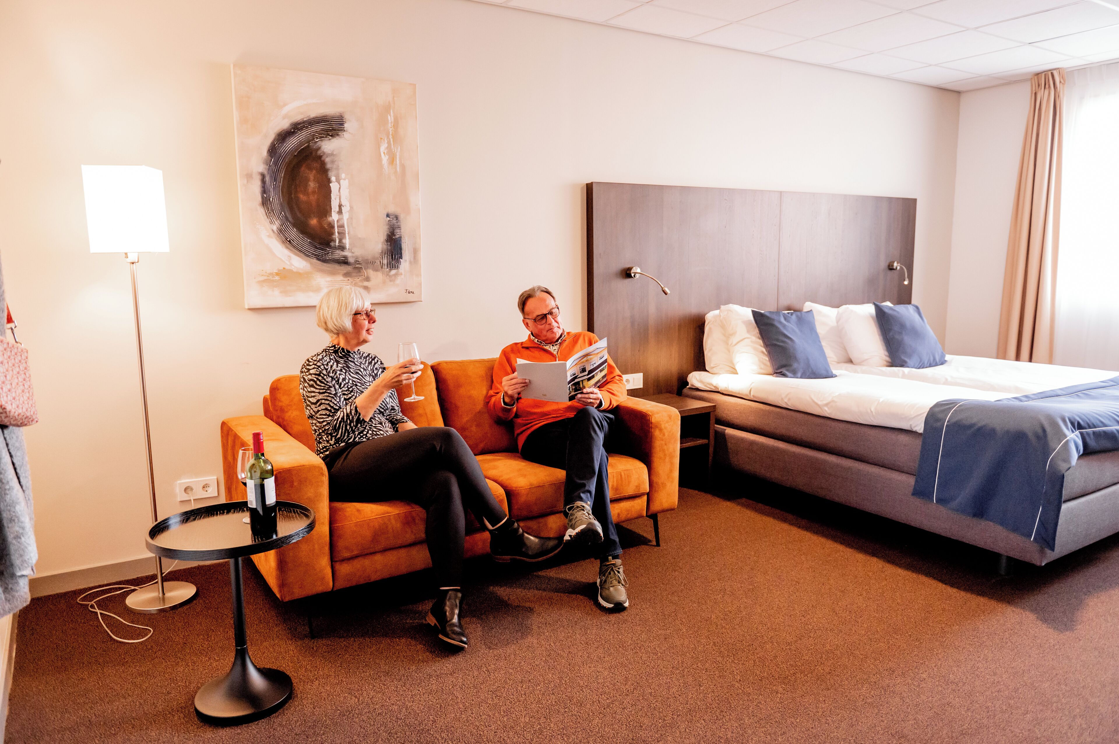 A hotel room with a bed, a sofa, a small table with a bottle, and a painting on the wall.