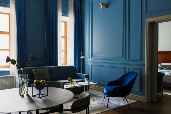 Une pièce avec un mur bleu, un canapé, un fauteuil bleu, une petite table avec un vase et des fruits, et une lampe.