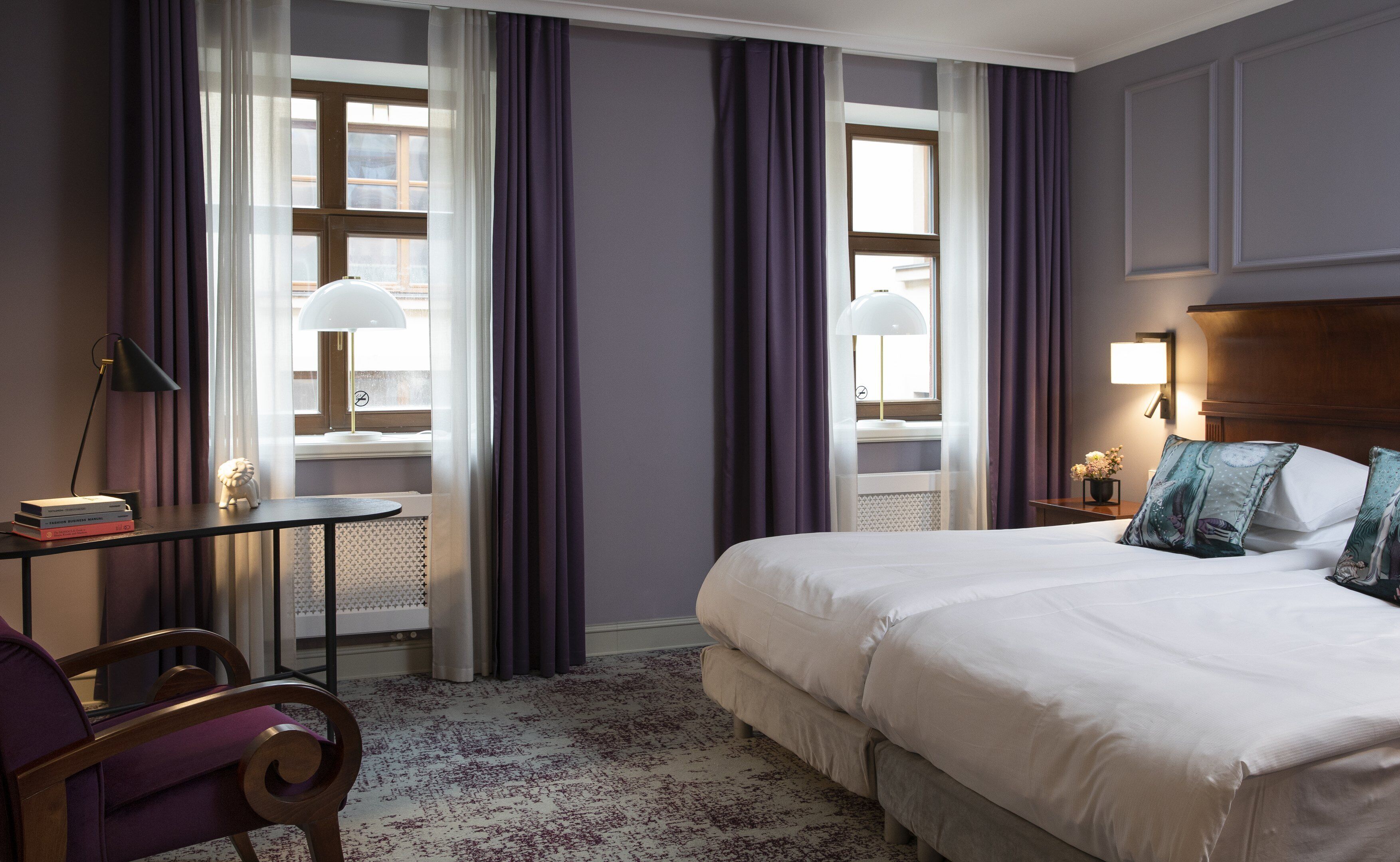 Une chambre d’hôtel avec un lit, un bureau, une chaise et deux fenêtres avec des rideaux.