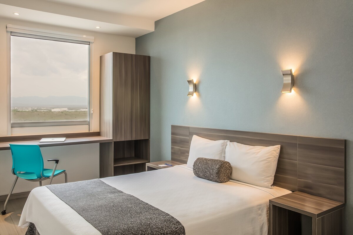 A modern hotel room with a large bed, a desk, and a chair. There is a window with a view of the landscape.