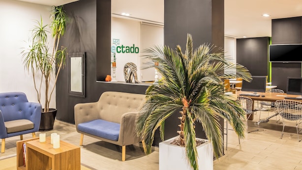 A modern reception area with a sofa, chairs, a coffee table, and potted plants. - One Monterrey Aeropuerto (Apodaca)