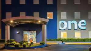 A hotel entrance with a 'one' sign, a covered entrance, and a landscaped area with bushes. - One Monterrey Aeropuerto (Apodaca)
