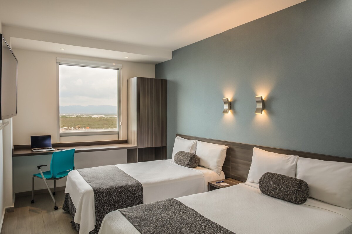 A hotel room with two beds, a desk with a laptop, a chair, and a large window offering a city view.