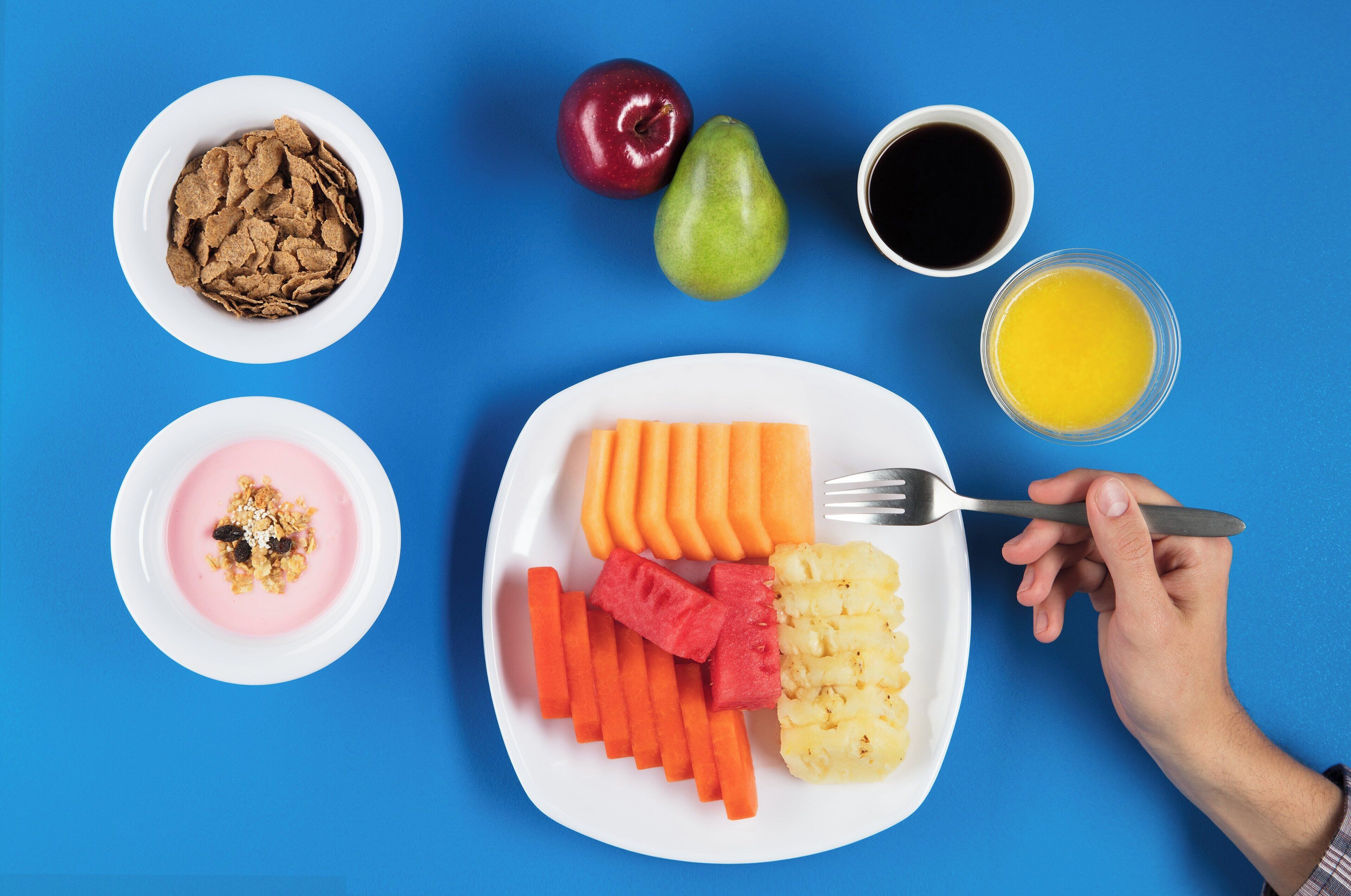 Une assiette contenant des carottes en tranches, de l’ananas et de la pastèque, un bol de céréales, une poire, une pomme et une tasse de café.