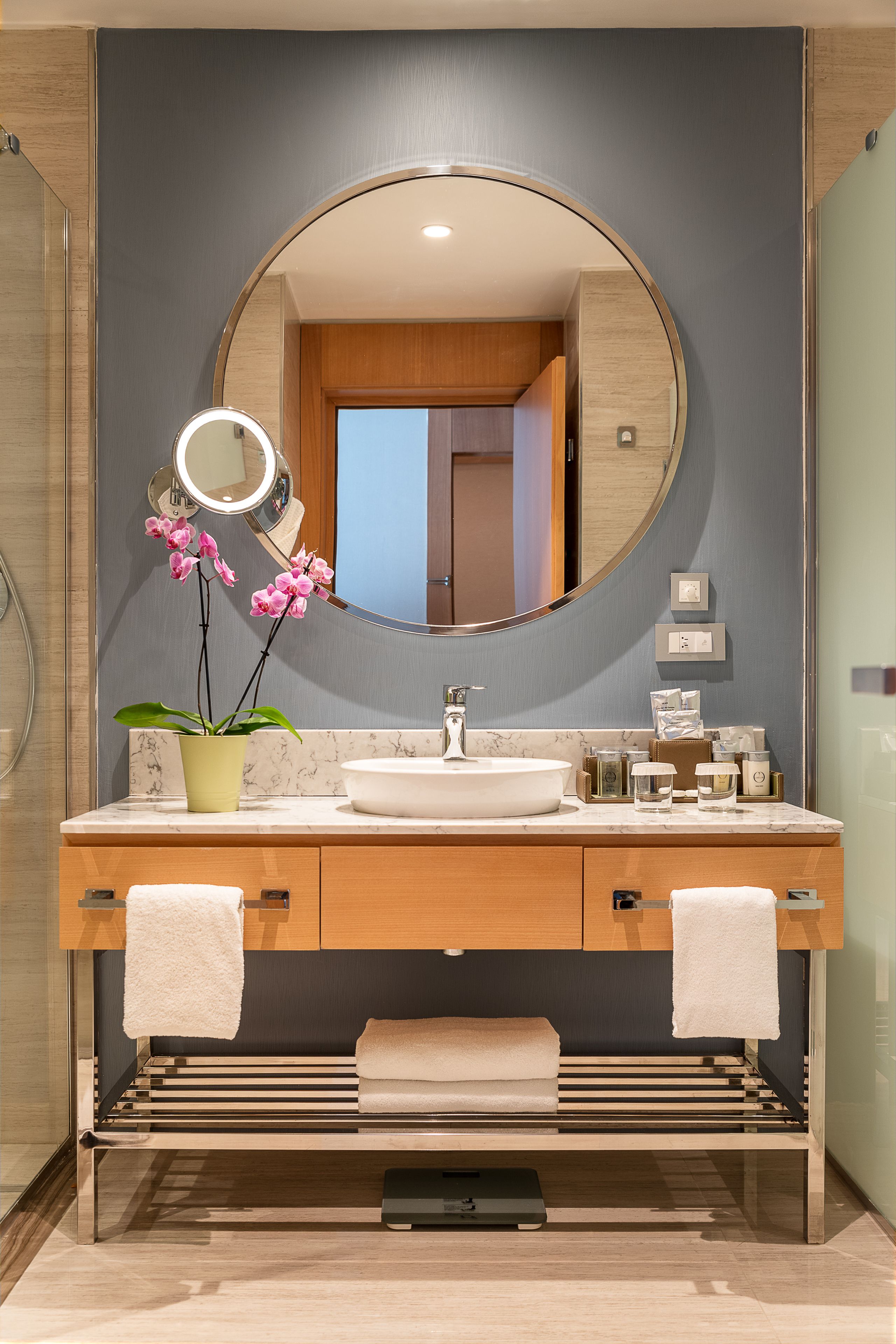 A modern bathroom with a round mirror, a sink, a potted orchid, and a towel rack.