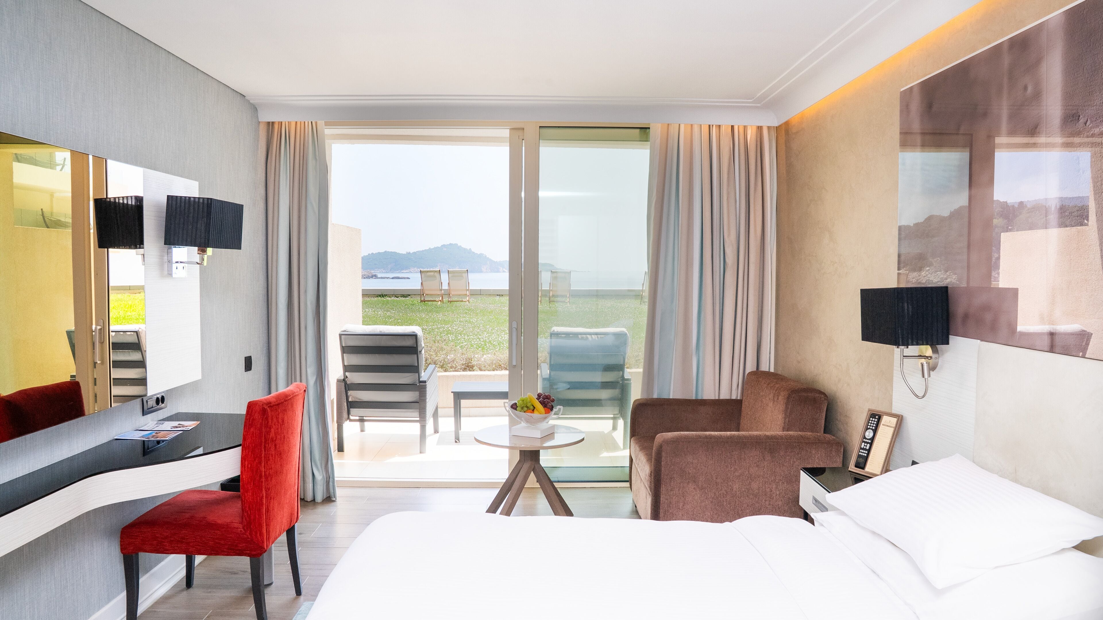 Ein Hotelzimmer mit Bett, Schreibtisch mit Stuhl, einer Couch, einem Fernseher und einem Balkon mit Meerblick.