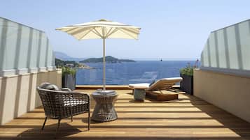 Um deck de madeira com vista para o mar, uma cadeira de vime, uma espreguiçadeira e um guarda-sol.