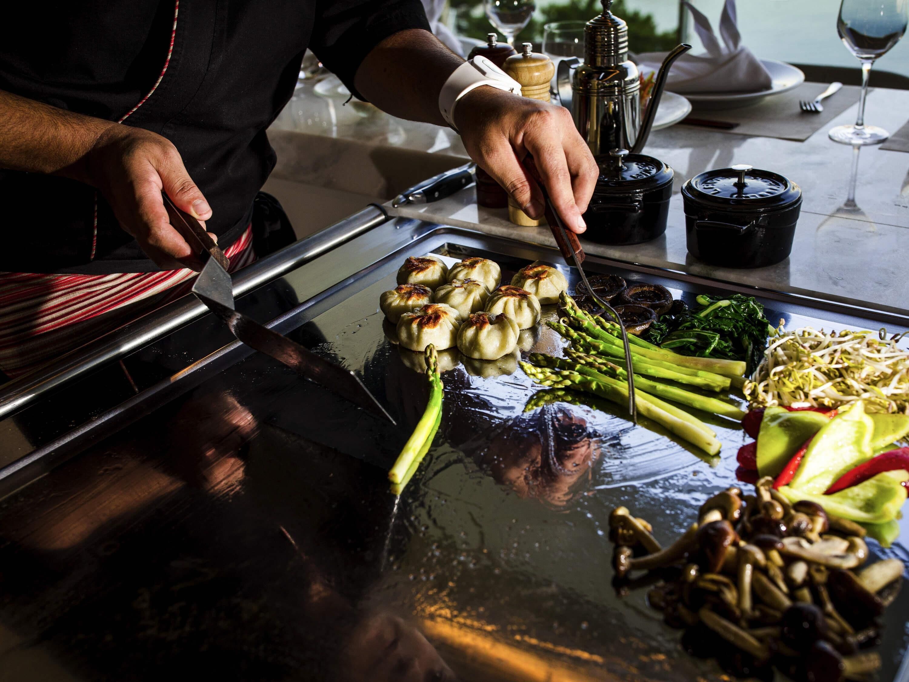 Un chef preparando comida en una parrilla abierta con verduras y albóndigas.