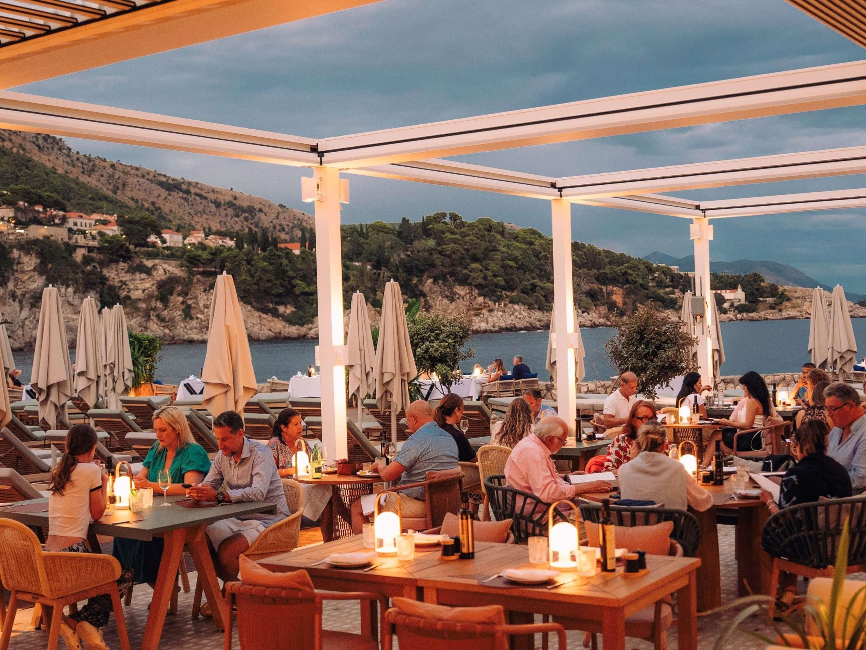An outdoor dining area with tables and chairs, umbrellas, and a view of the sea and hills.