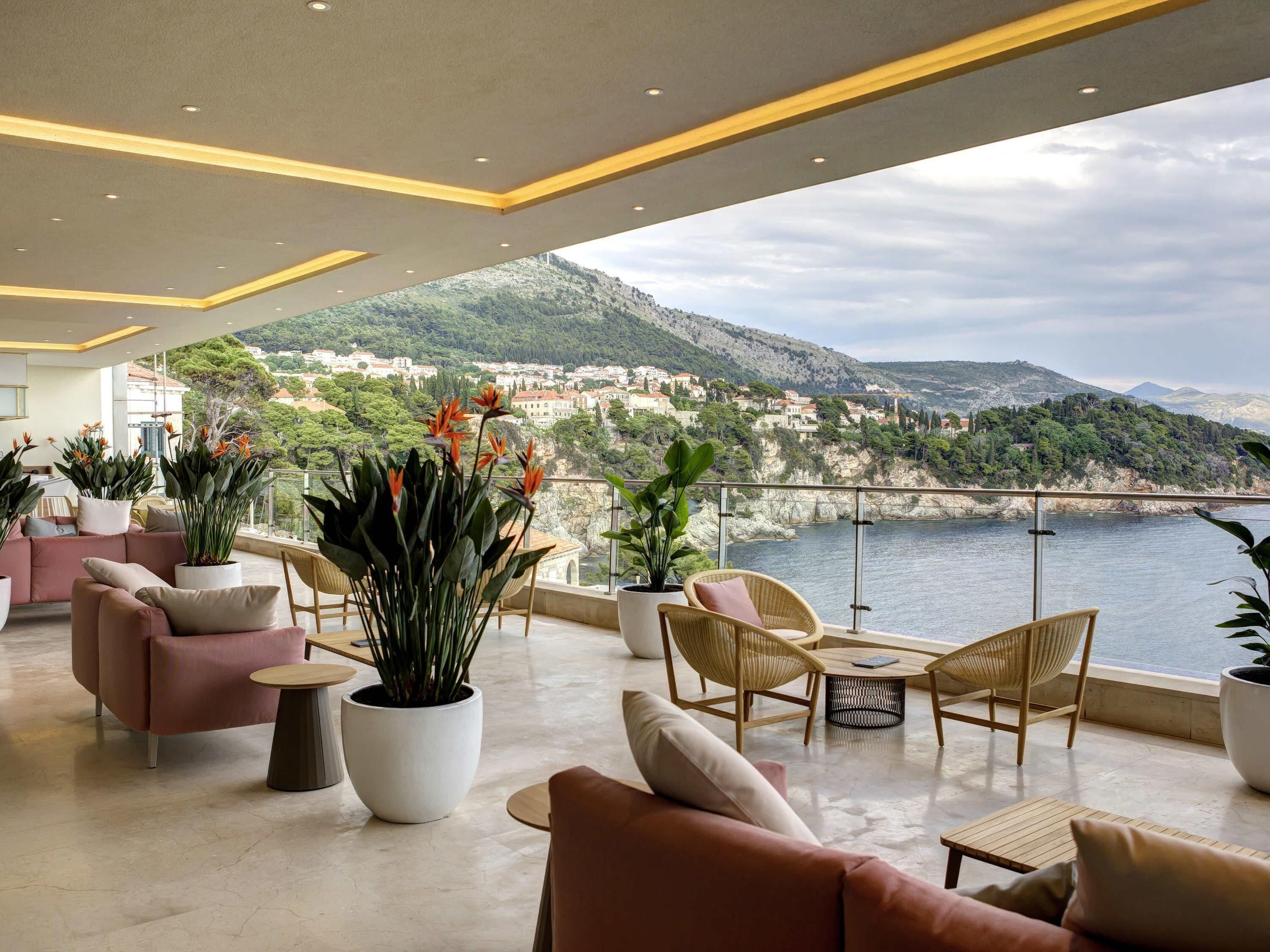 A spacious balcony with modern furniture, potted plants, and a scenic view of a coastal town and mountains.