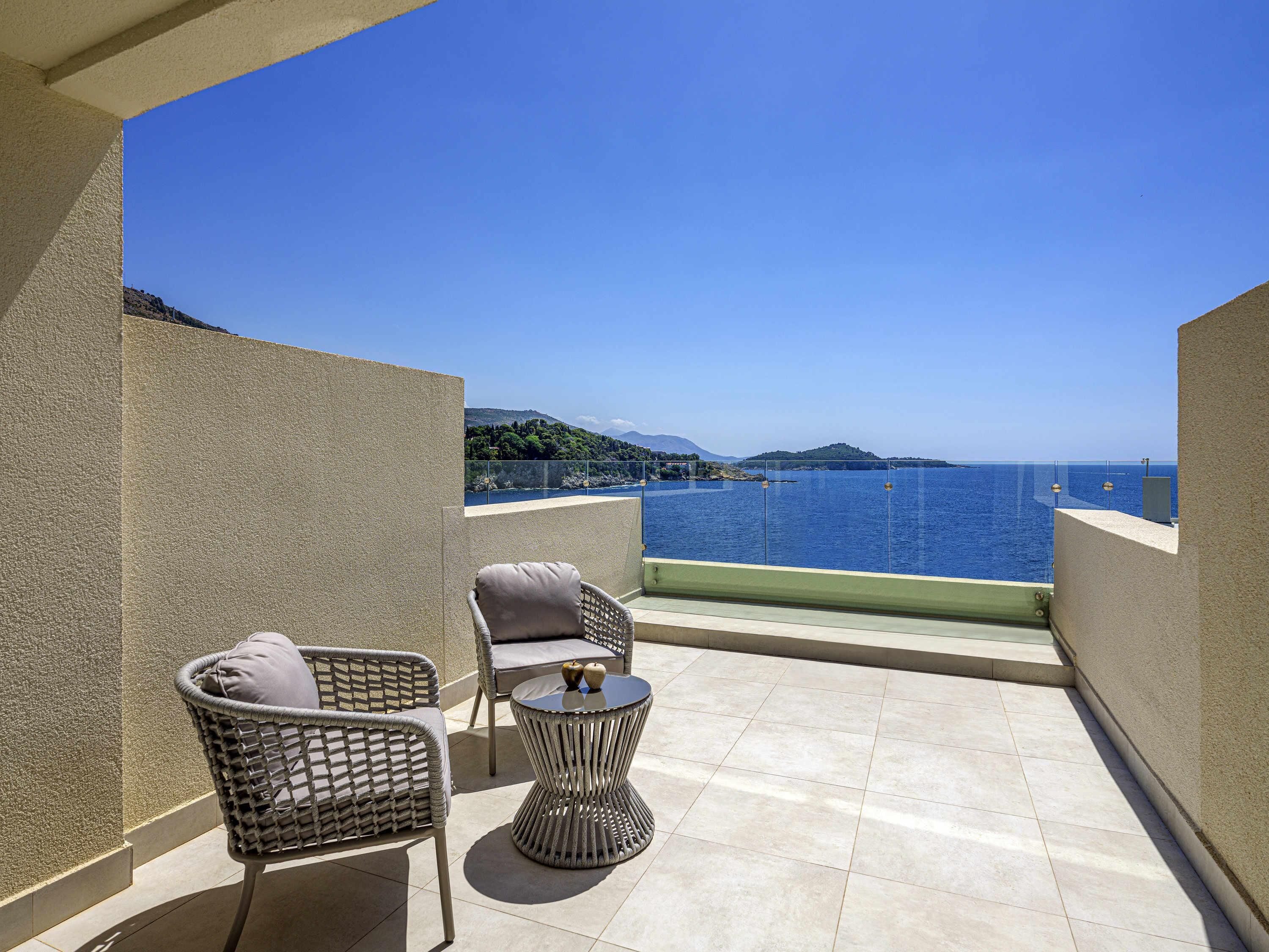 A balcony with two wicker chairs and a small round table, overlooking a clear blue sea and distant hills.