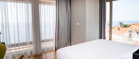 A hotel room with a large bed, a view of the sea, and a balcony with a view of the sea and buildings.