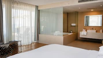 A modern hotel room with a large bed, a glass-enclosed bathroom, and a view of the outdoors.