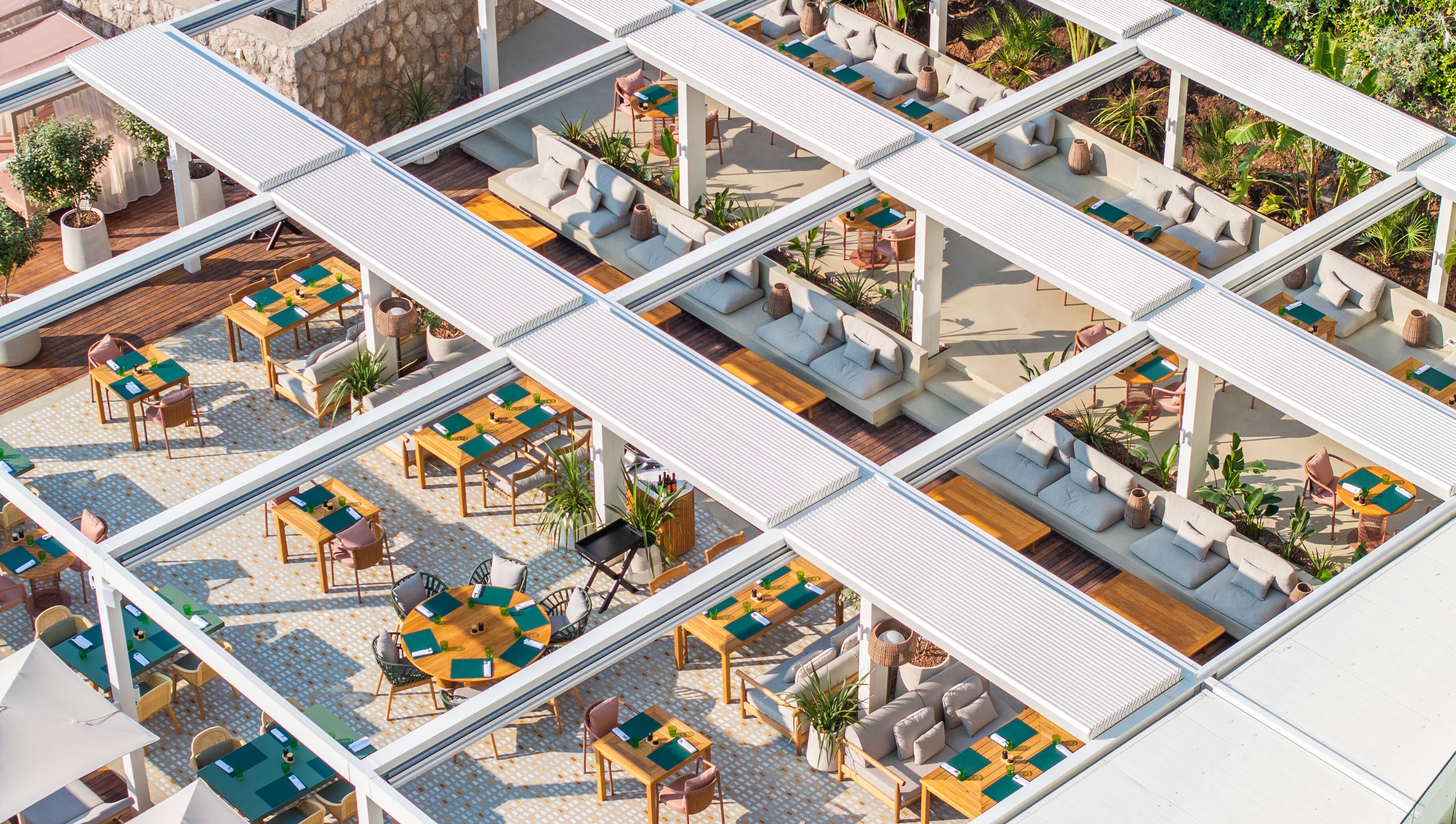 Una terraza en la azotea con varias zonas de estar, incluyendo sofás y mesas, rodeada de macetas con plantas y un muro de piedra.