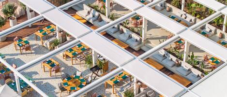 Una terraza en la azotea con varias zonas de estar, incluyendo sofás y mesas, rodeada de macetas con plantas y un muro de piedra.