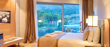 Ein Hotelzimmer mit einem großen Bett, einem Schreibtisch mit Stuhl, einem Fernseher und einem Fenster mit Blick auf eine gebirgige Landschaft.