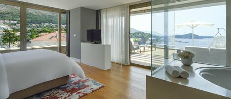 A modern hotel room with a large bed, a television, a balcony with a view of the sea, and a bathroom with a bathtub.