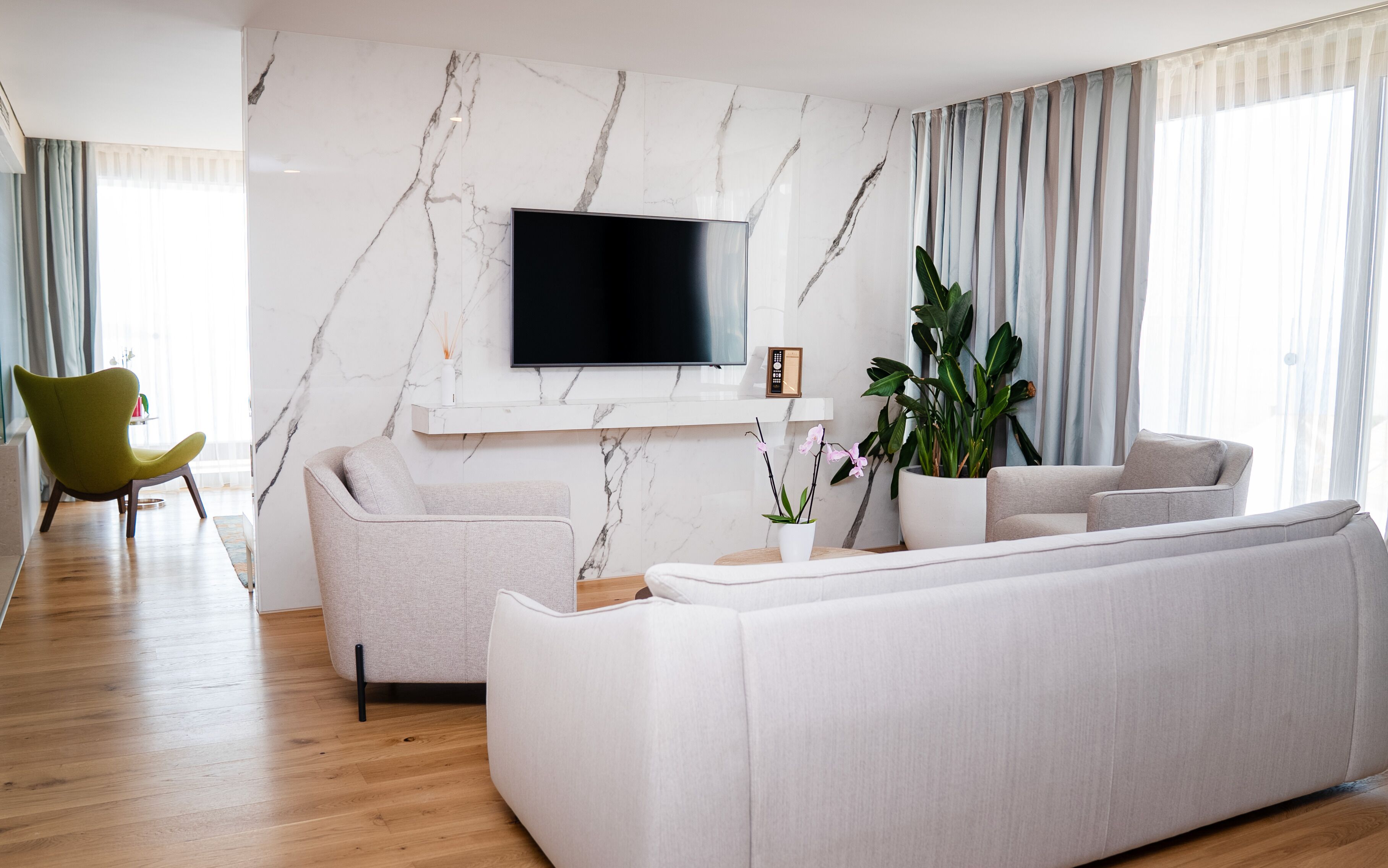 A modern living room with a marble wall, a flat-screen TV, a grey sofa, a green armchair, and a large potted plant.
