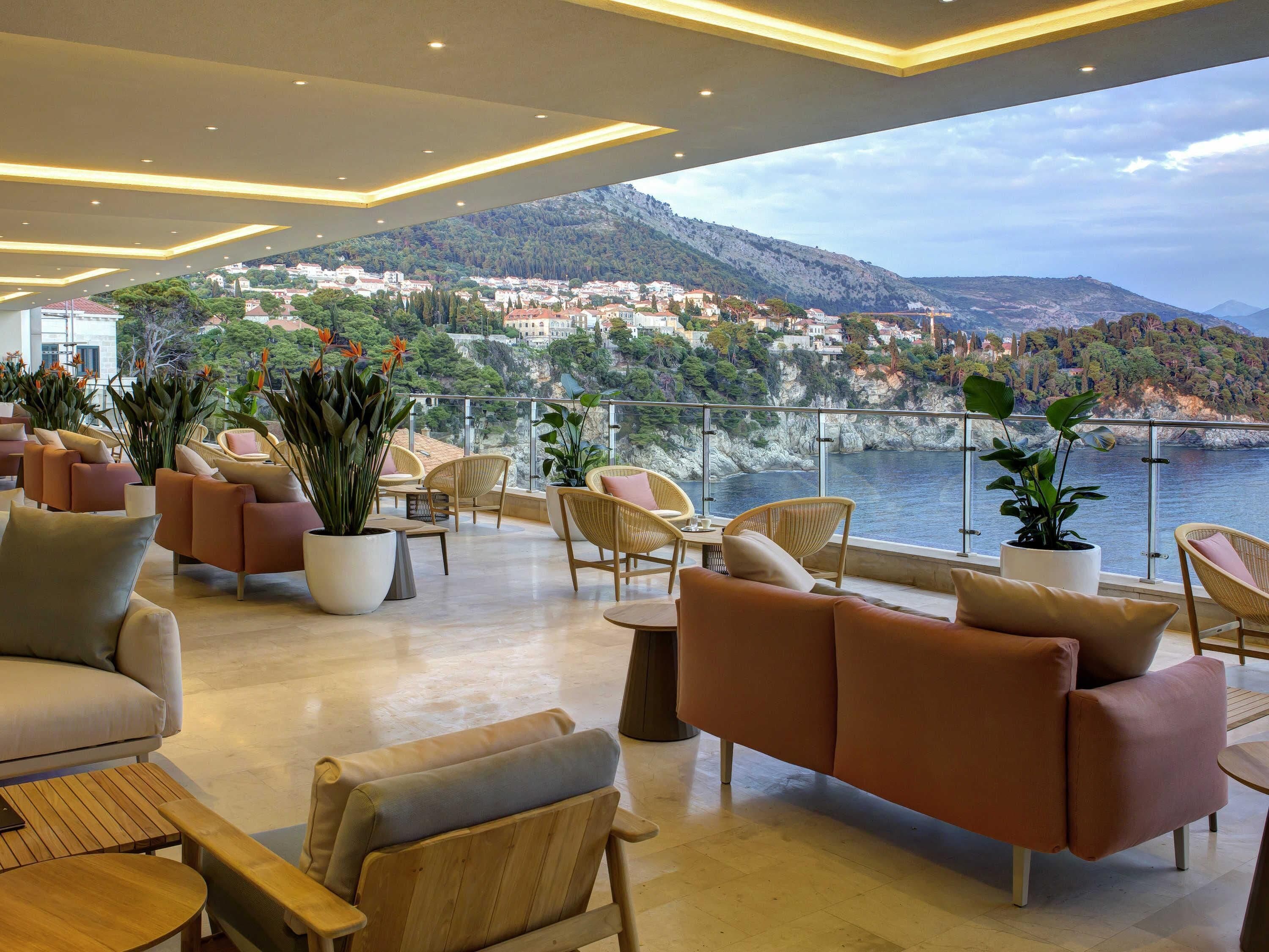 A modern lounge with comfortable seating and potted plants, overlooking a coastal town and mountains.