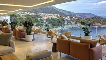 Un salon moderne doté de sièges confortables et de plantes en pot, offrant une vue sur une ville côtière et les montagnes.
