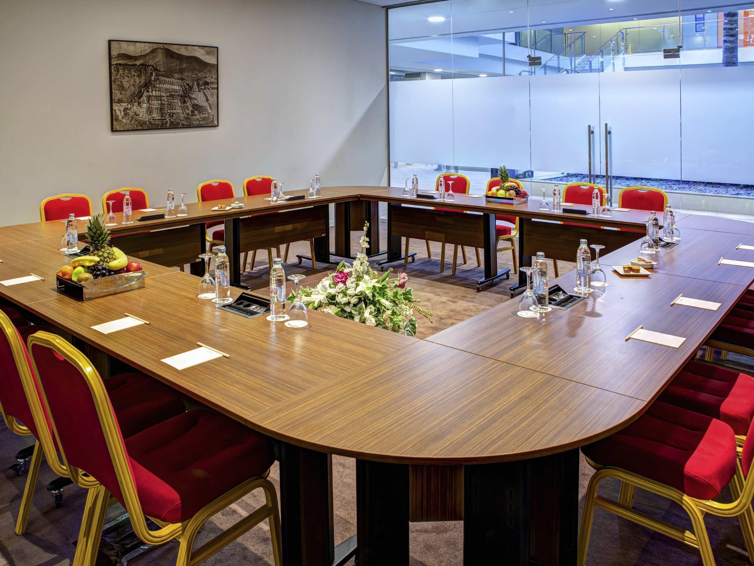 A conference room with a long table, red chairs, water bottles, and a floral centerpiece.