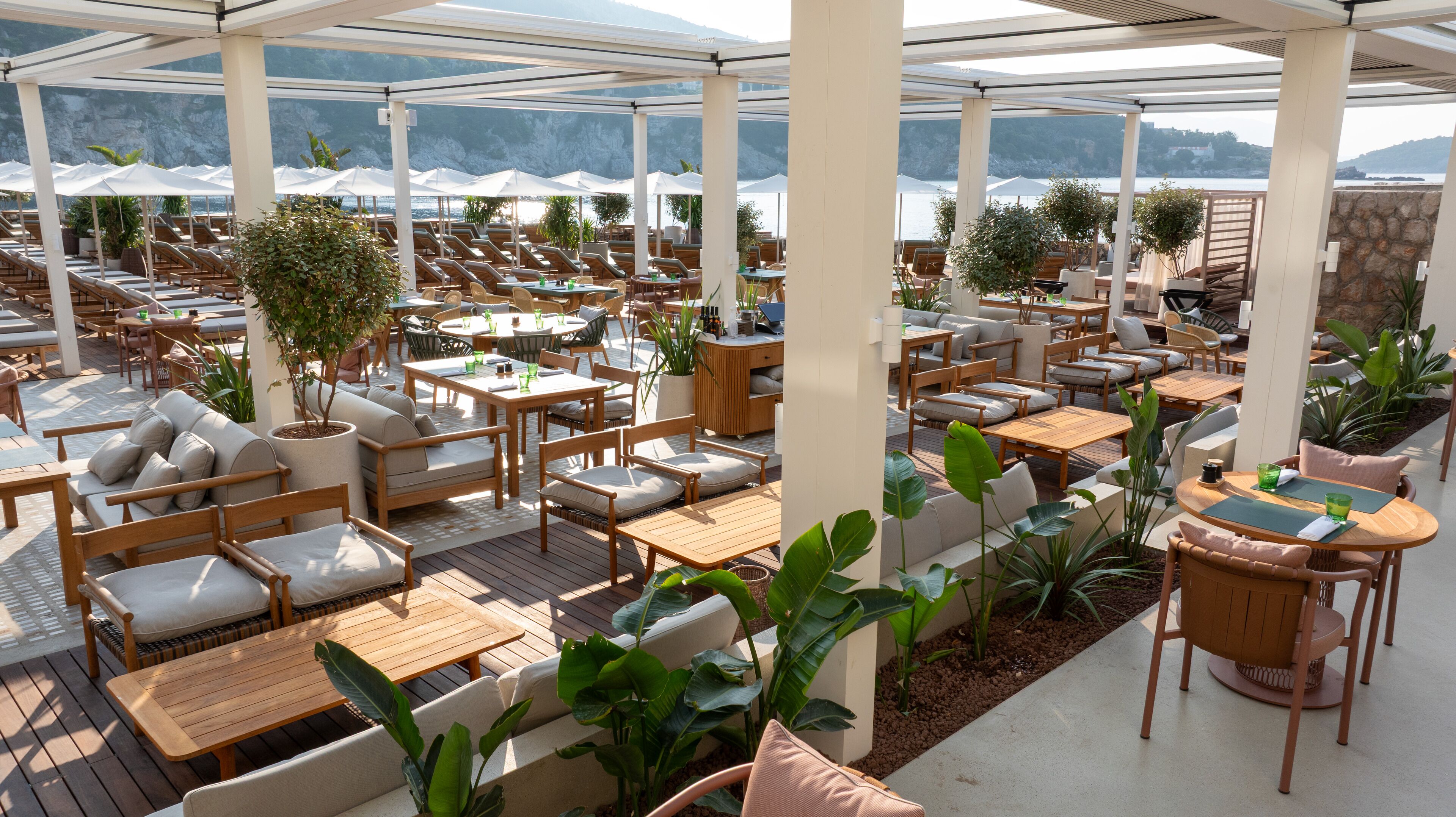 A covered outdoor seating area with multiple tables and chairs, surrounded by potted plants and overlooking a body of water.
