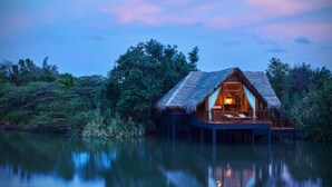 A thatched-roof bungalow on stilts over water, with a visible interior and a reflection in the calm water. - Jetwing Vil Uyana, A Luxury Reserve (Sigiriya)