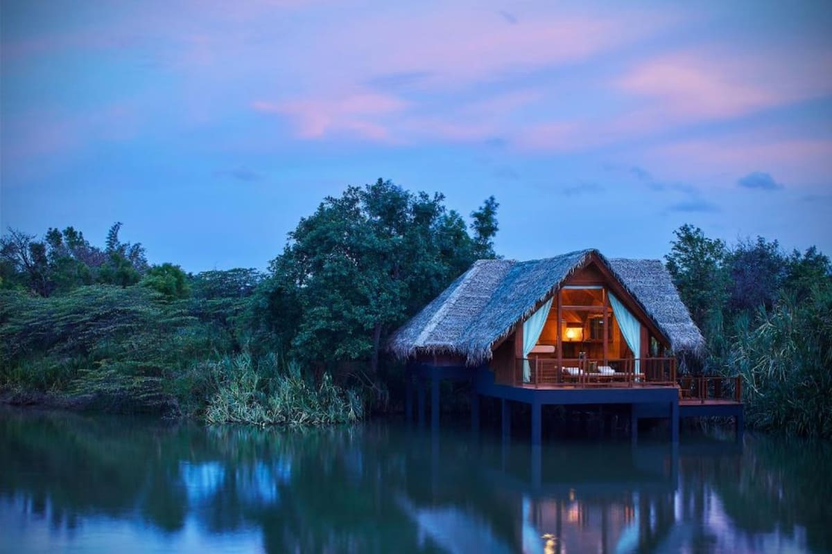A thatched-roof bungalow on stilts over water, with a visible interior and a reflection in the calm water.