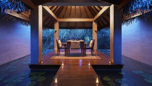 An indoor dining area with a wooden table set for two, surrounded by chairs, with a view of a pond and foliage outside. - Jetwing Vil Uyana, A Luxury Reserve (Sigiriya)