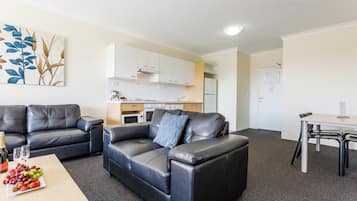 Ett modernt vardagsrum med en svart lädersoffa, ett träbord med frukt och en flaska, och ett köksområde i bakgrunden.