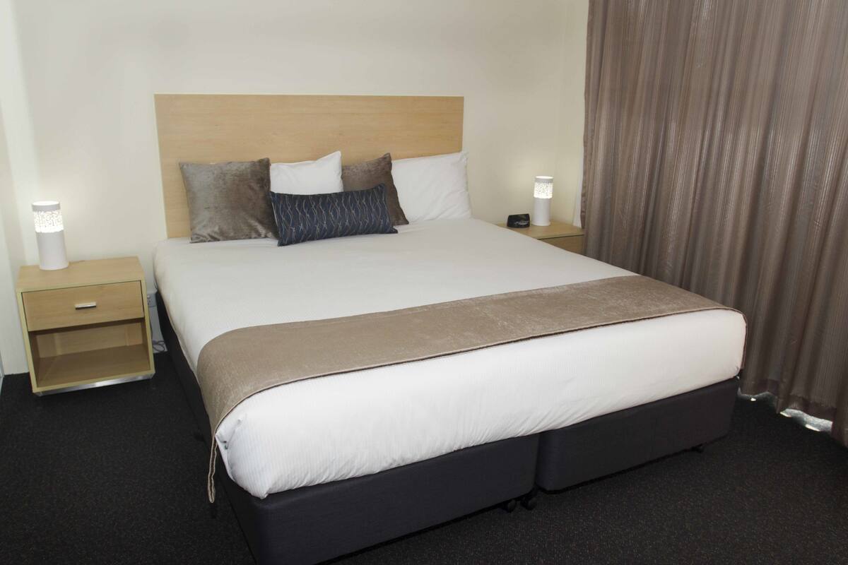 A hotel room with a large bed, two bedside tables, a nightstand, and a curtain.