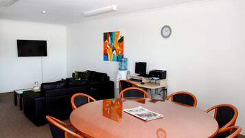 Een moderne vergaderruimte met een glazen tafel, oranje stoelen, een zwarte bank, een flatscreen-televisie, een klok en een kleurrijk abstract schilderij.